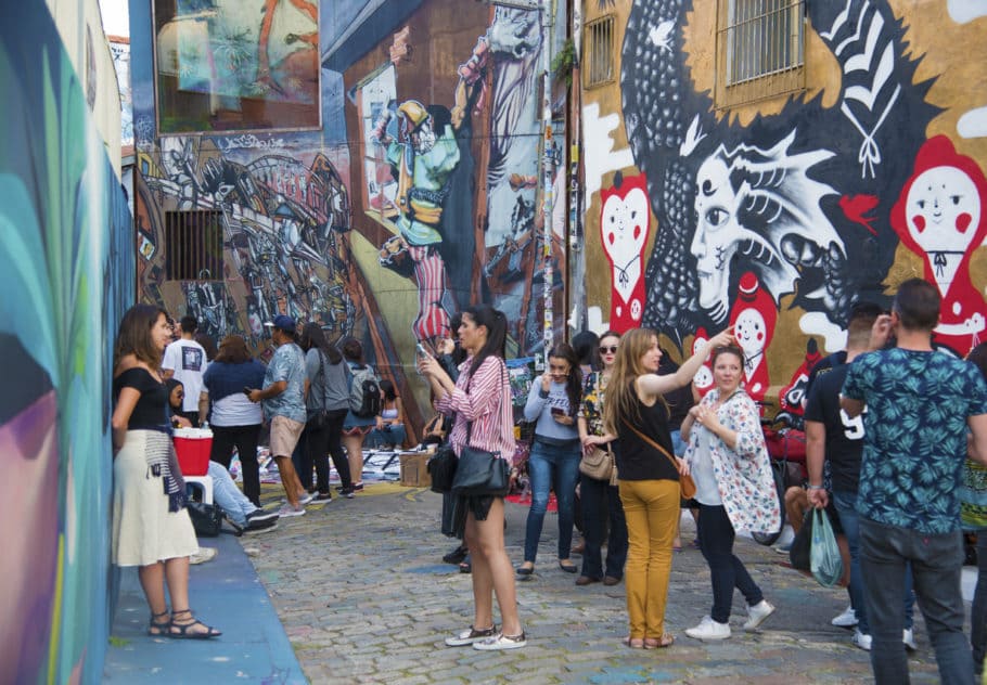 Rua com muros altos e repletos de pinturas. Há pessoas na rua entre os muros observando os desenhos e tirando fotos. Local: Beco do Batman em São Paulo